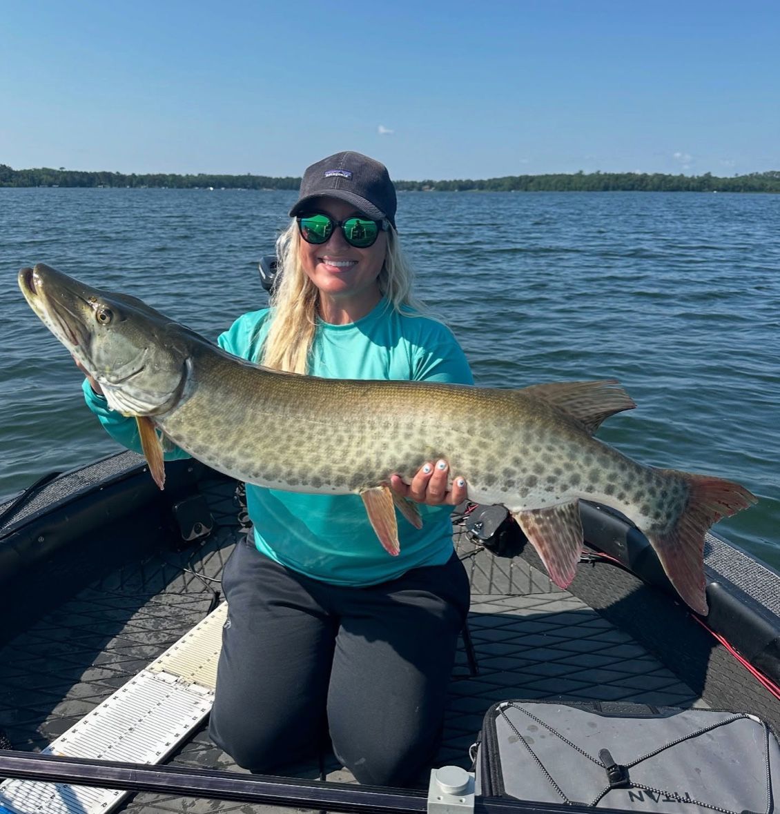 Woman on boat holds large muskie fish; blue sky and water.