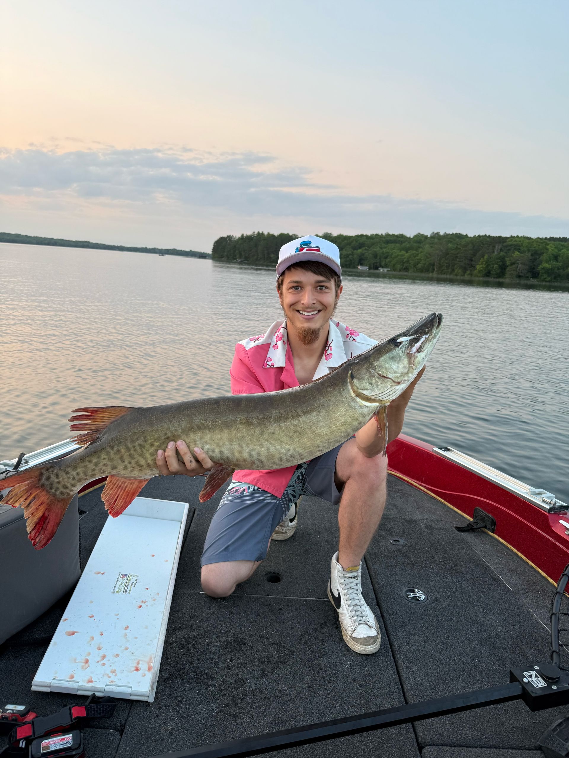 Teenager kneels on a boat, holding a large fish. Lake in the background.