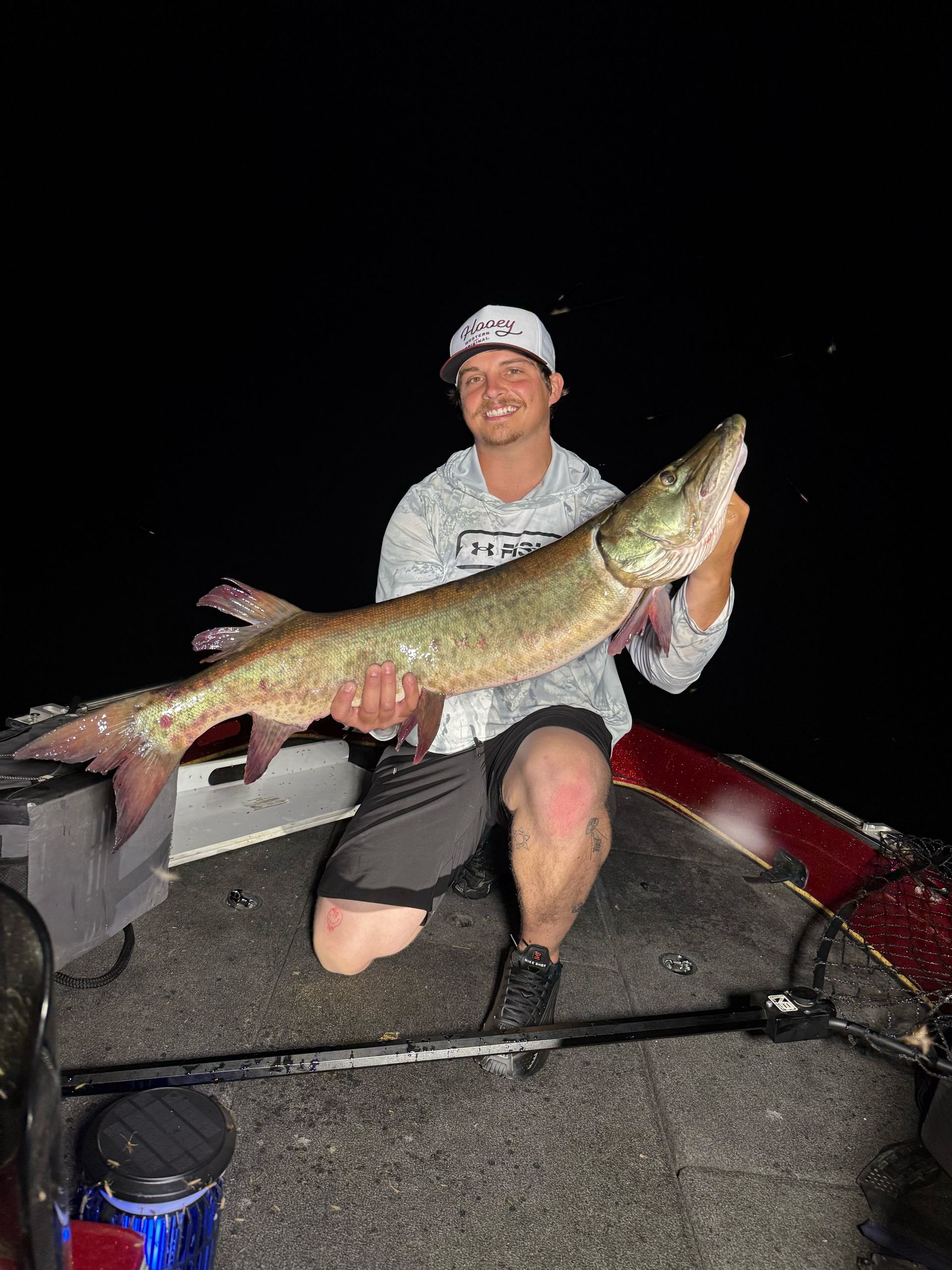 Man kneeling on boat holding a large fish, presumably a muskie, against a dark background.