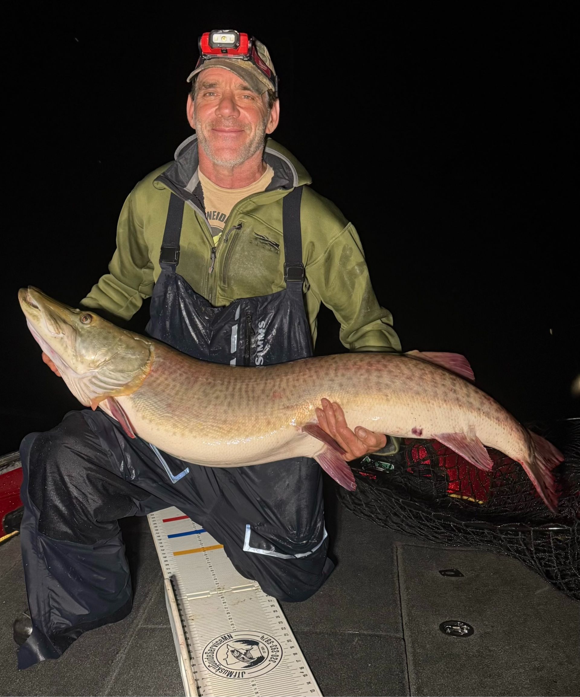 Man holding a large muskie fish on a boat at night. He is wearing a headlamp, waders, and smiling.