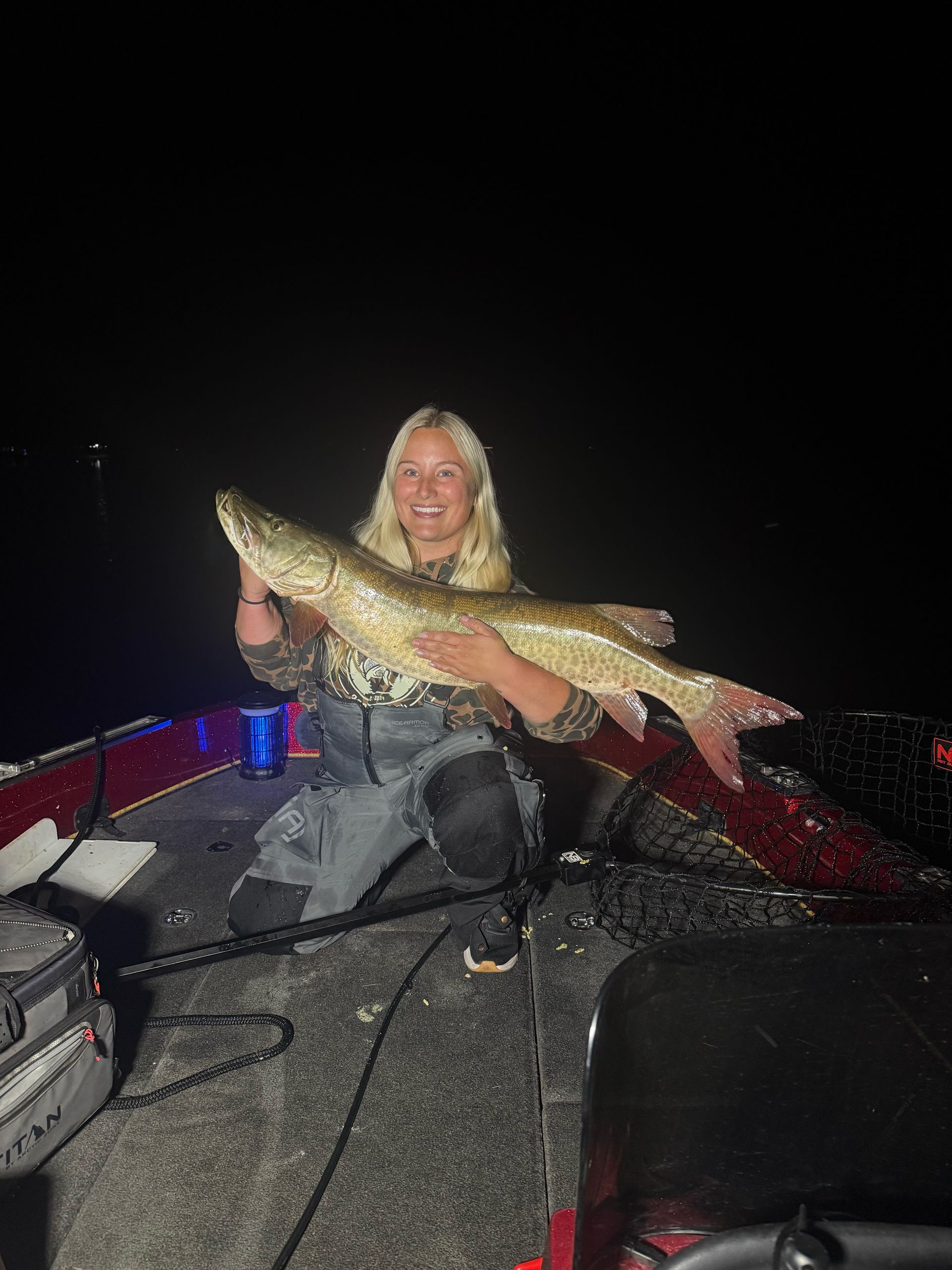 Woman in camouflage holds a large fish in a boat at night.