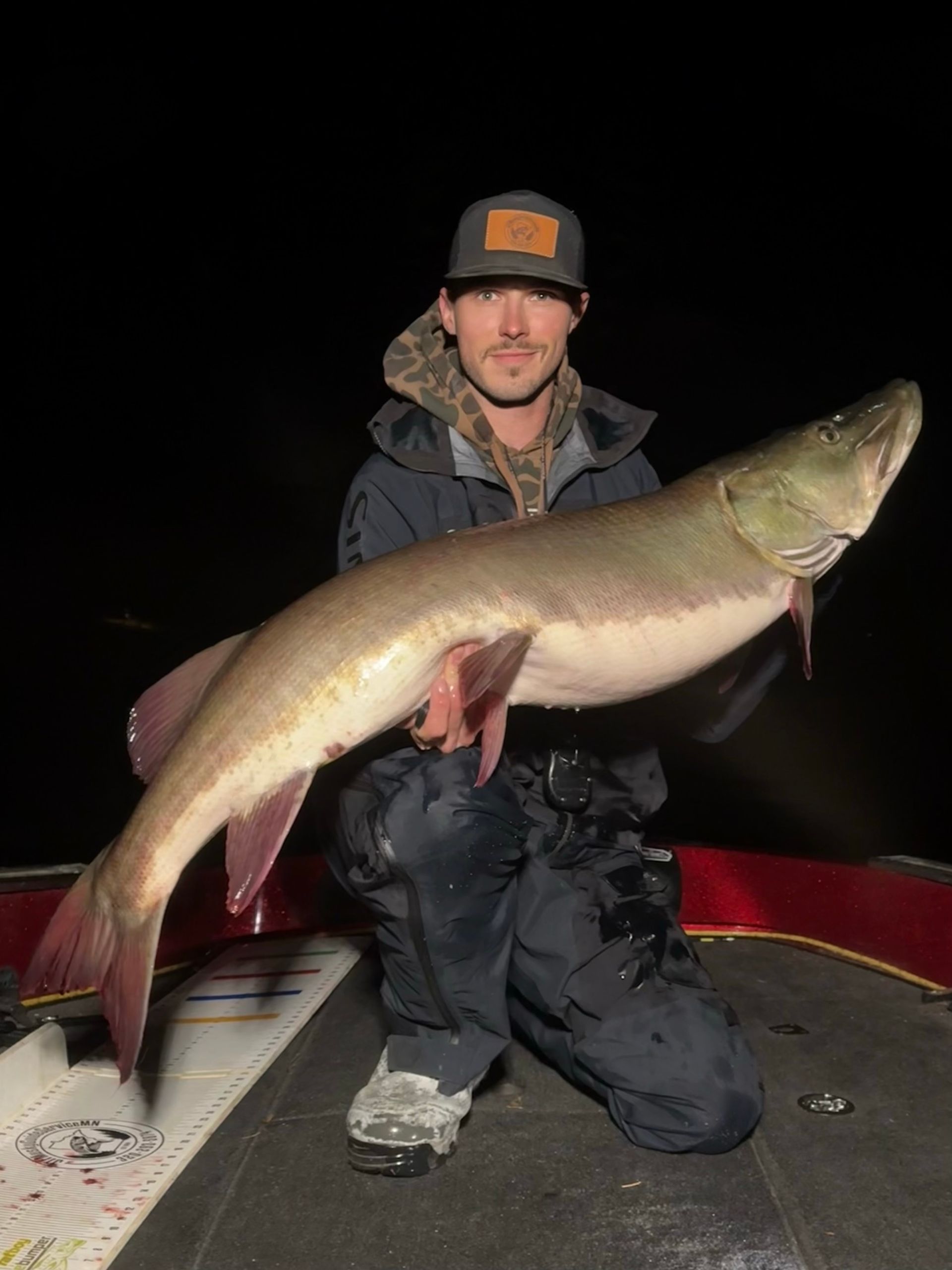 Man kneels on boat, holding a large muskellunge. Dark setting.