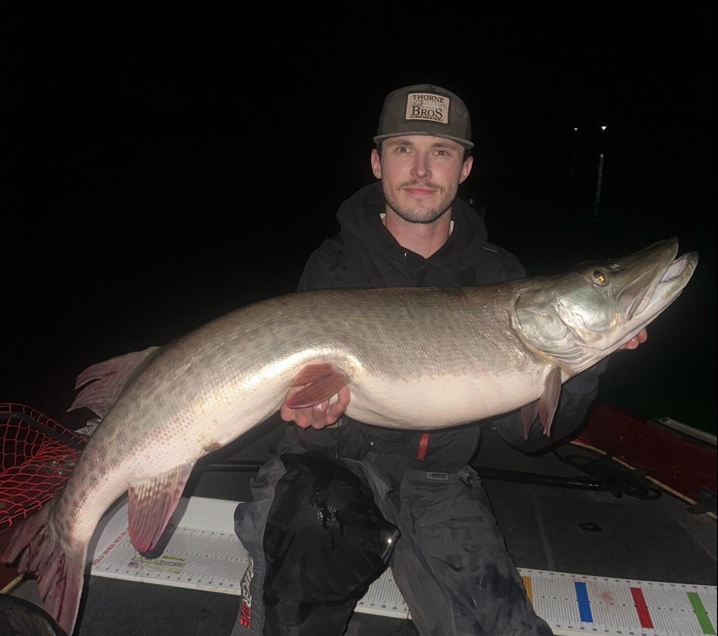 Man holding a large muskie fish, caught at night. The fish is light-colored, and the man wears a hat and dark clothing.