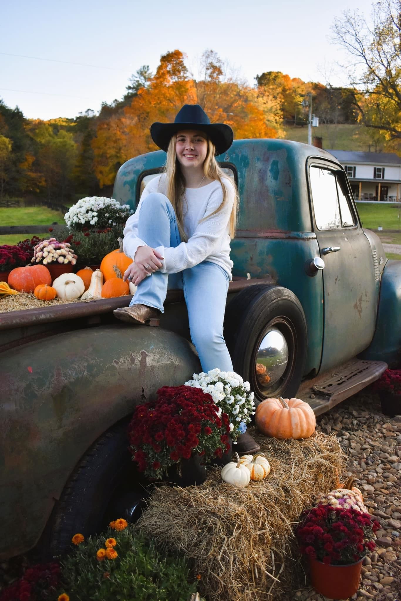 A woman is sitting on the back of an old truck surrounded by pumpkins and flowers.
