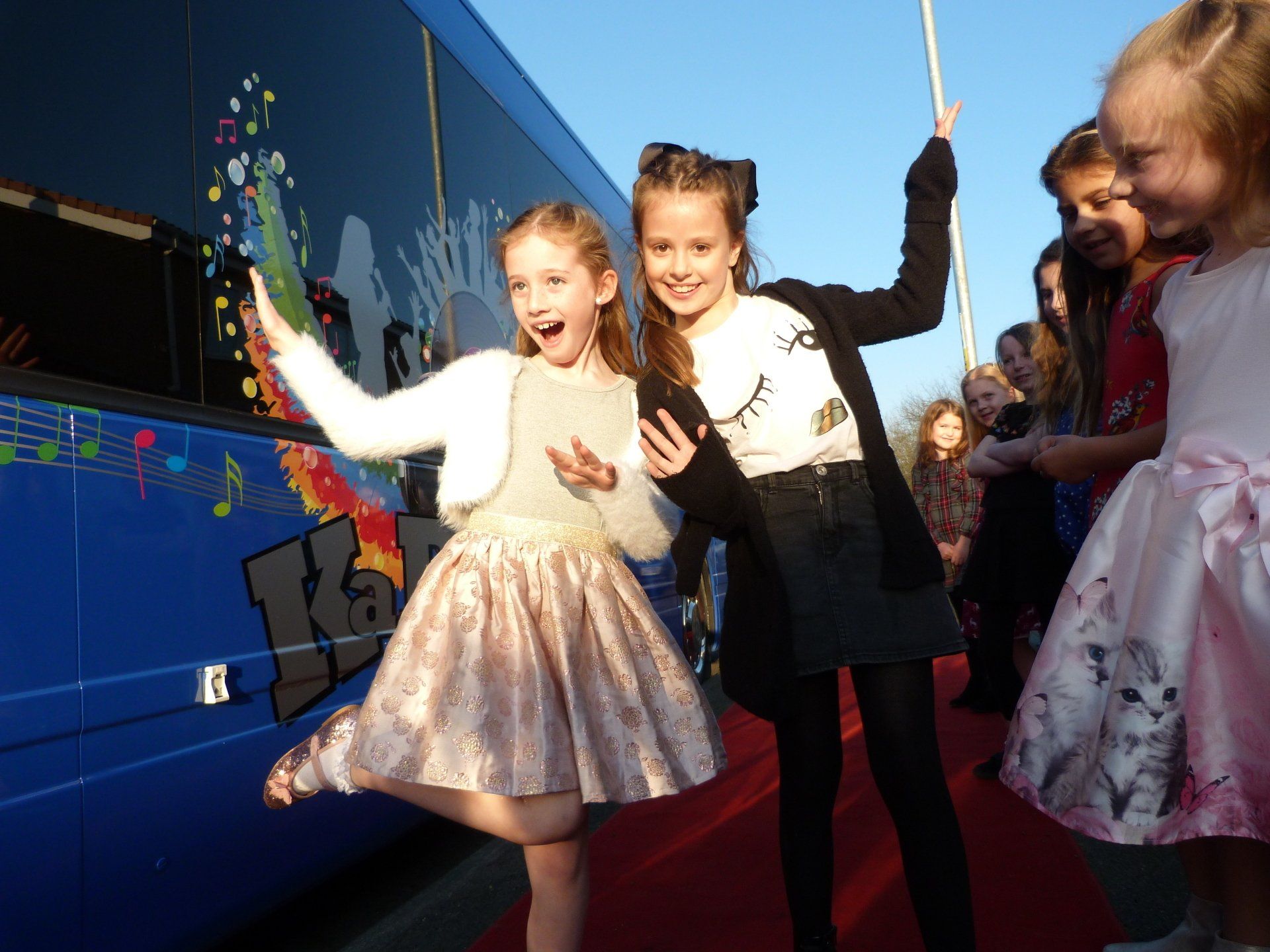 Two little girls are standing on a red carpet in front of a blue bus.