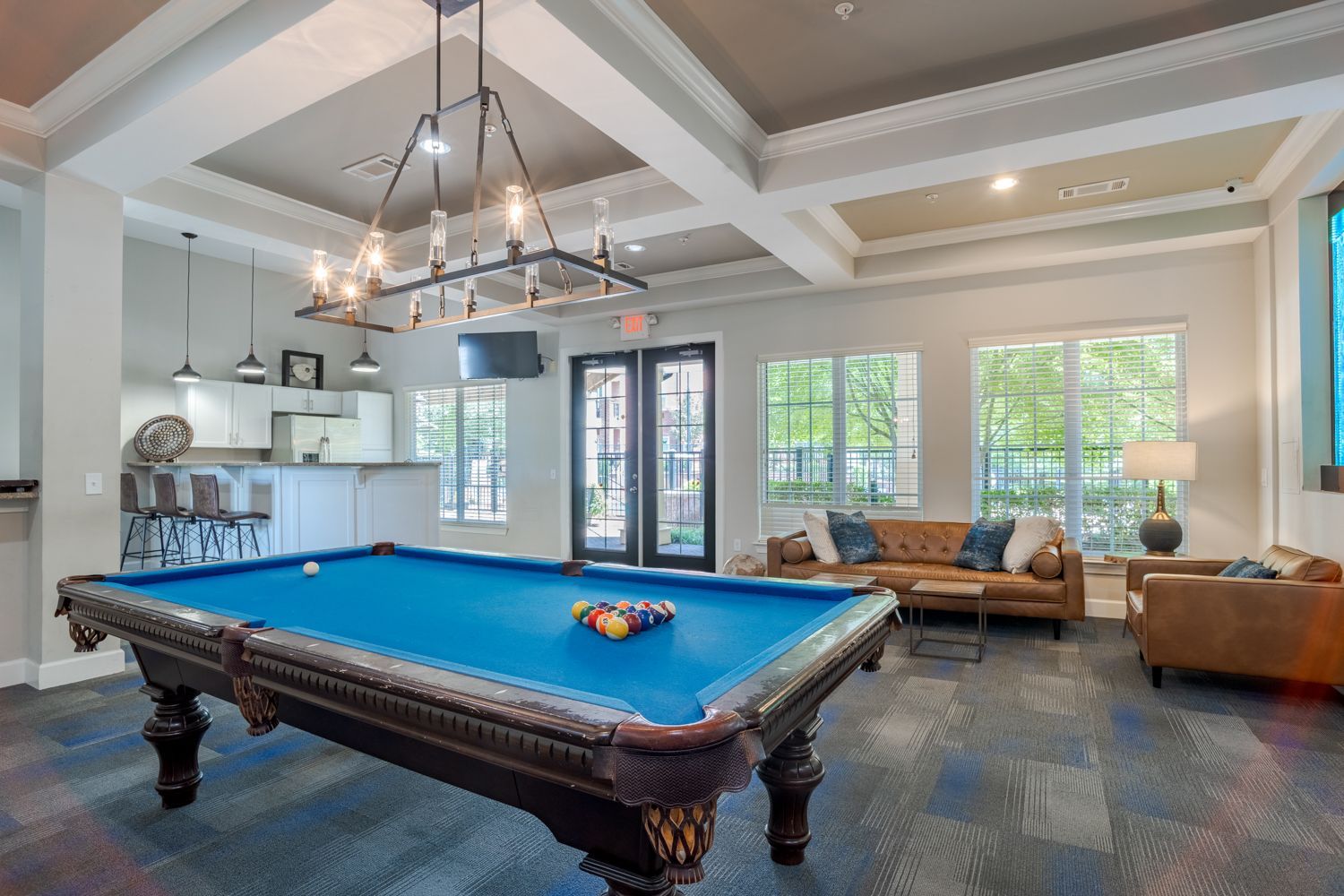 Pool table in a common area with seating, doors, windows, and a kitchen visible.