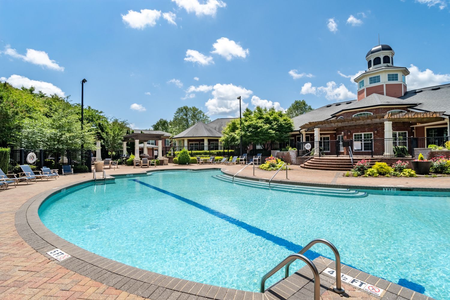 Swimming pool with brick edging, lounge chairs, and a clubhouse under a blue sky.