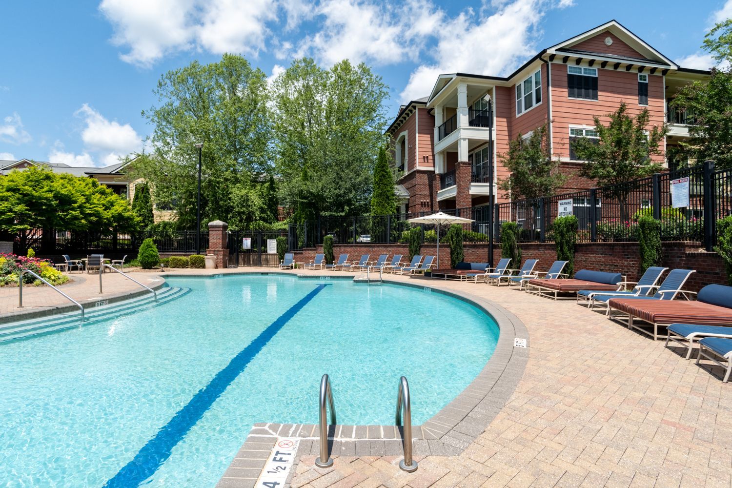 Swimming pool with lounge chairs, brick building in background. Sunny day.