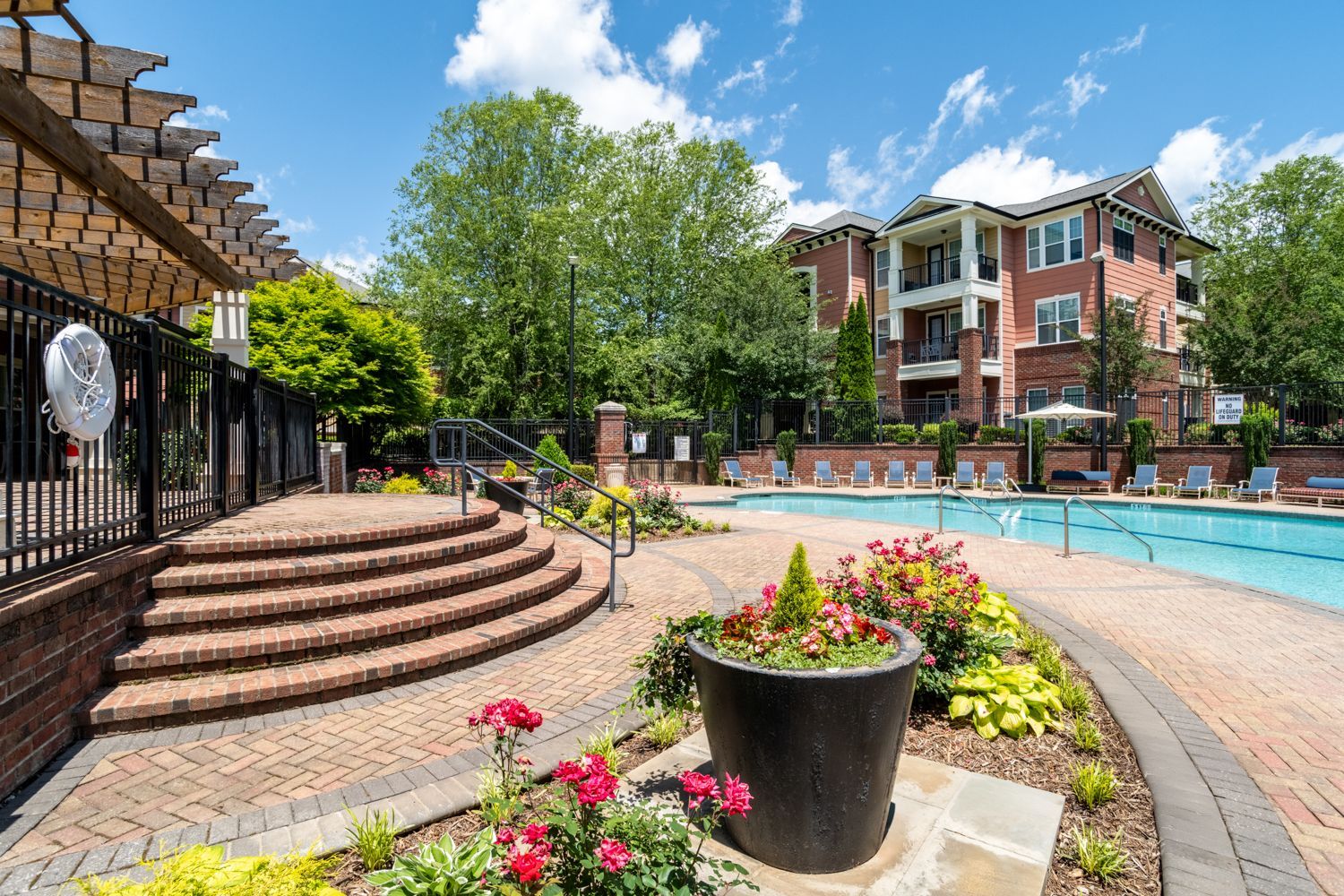 A brick patio leads to a swimming pool at an apartment complex under a sunny sky.