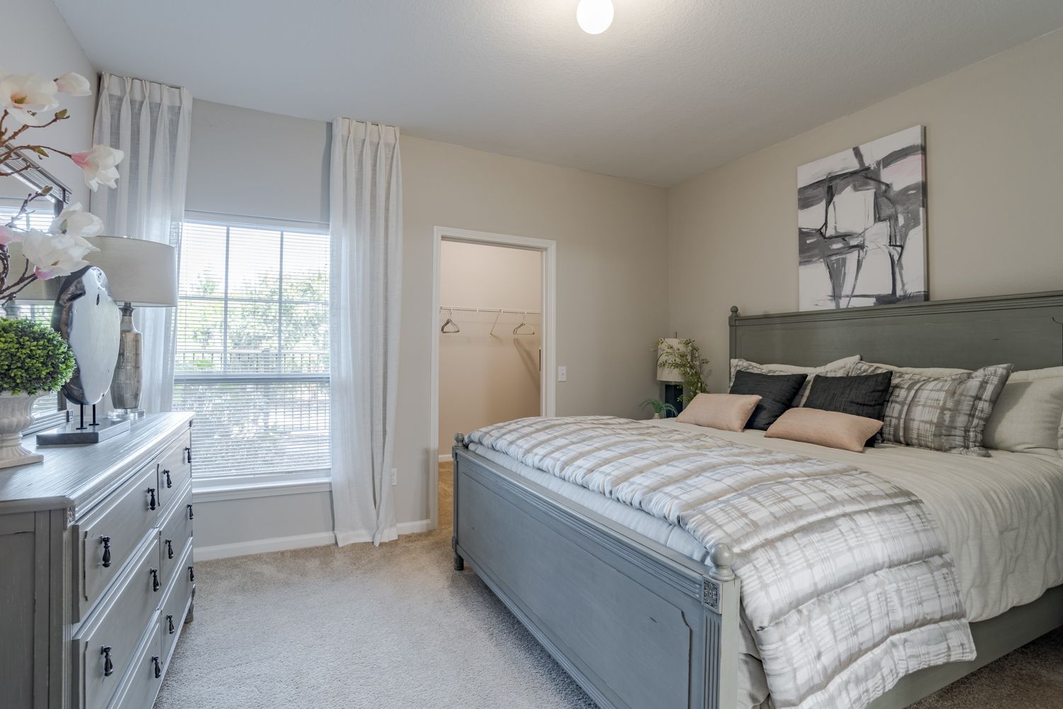 Bedroom with a gray bed, dresser, and wall art. White curtains and neutral walls.