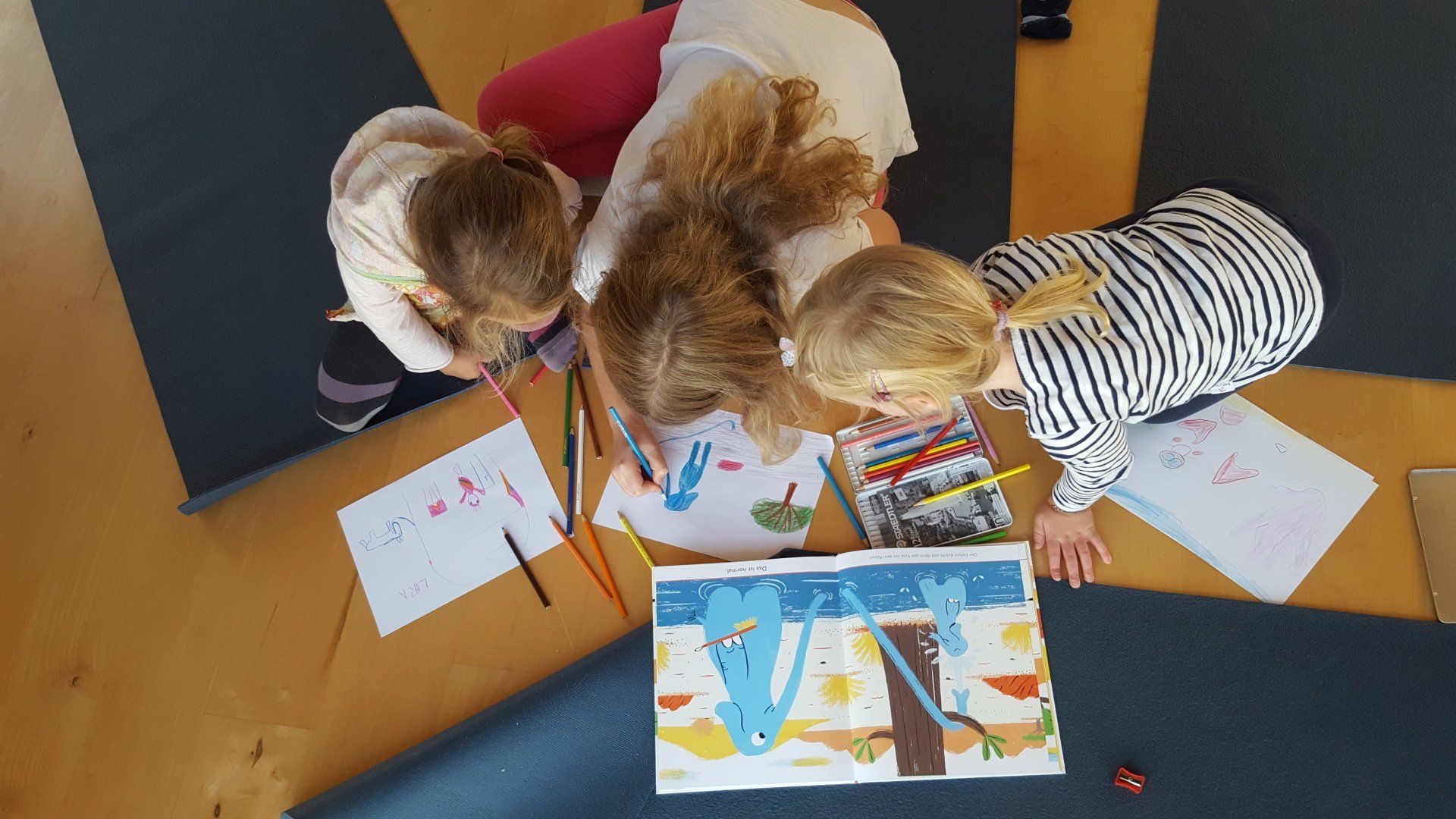 Drei junge Mädchen sitzen auf einer Yogamatte und zeichnen auf einem Blatt Papier.