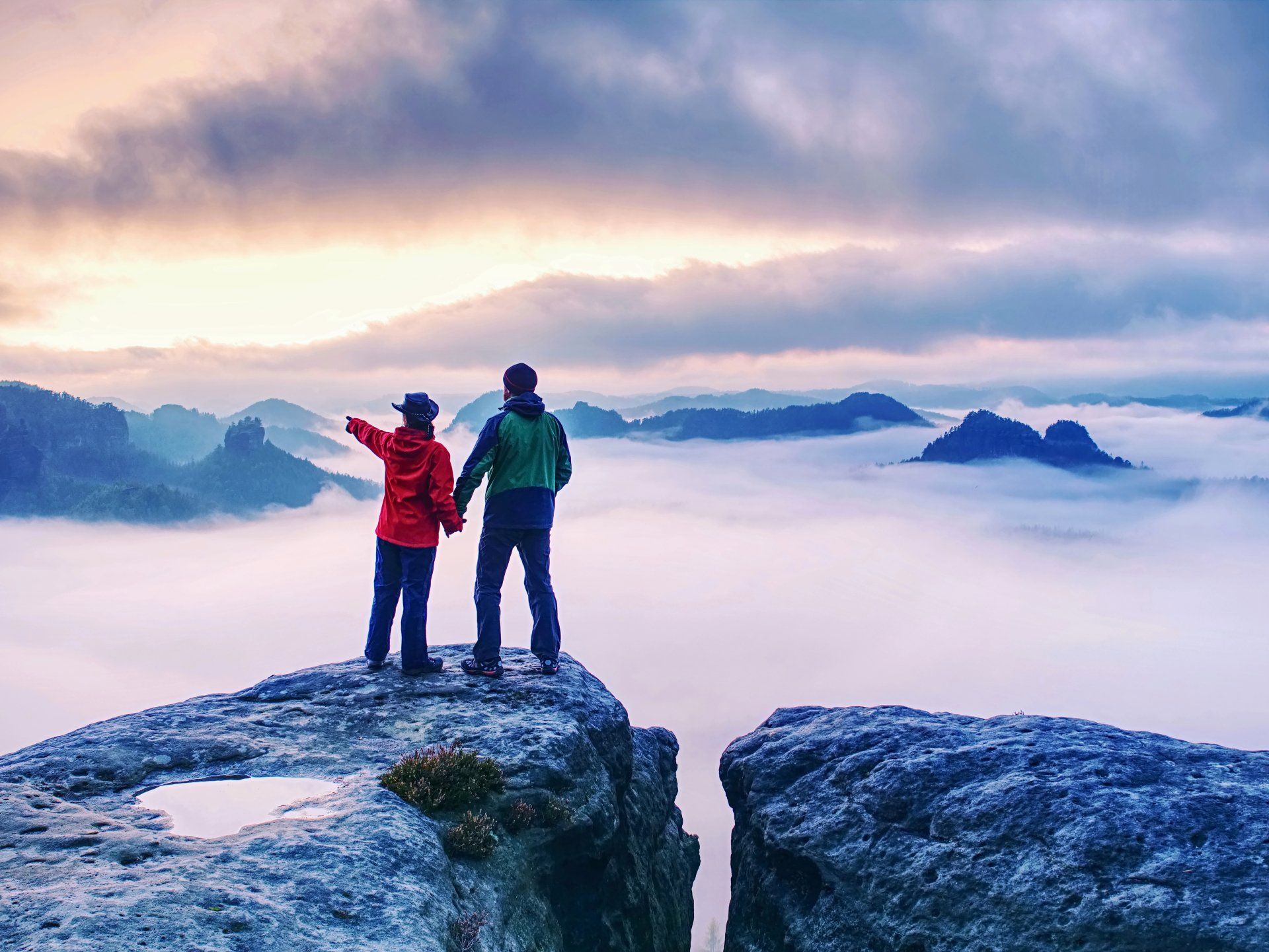 Zwei Menschen stehen am Rand einer Klippe und blicken auf ein wolkenverhangenes Tal.