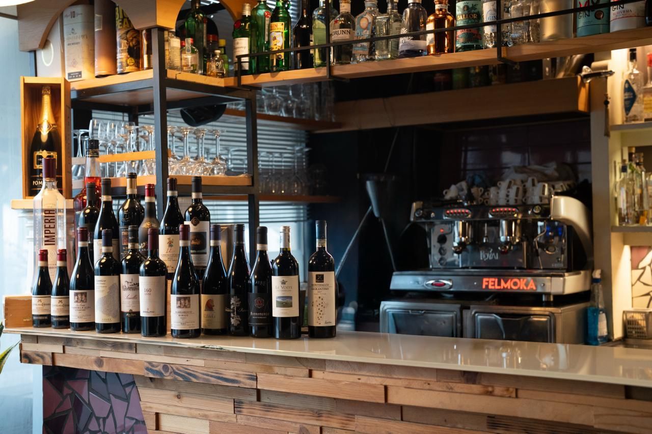 Bar all'aperto con tavoli, sedie e vetrate. Alberi in primo piano, luci e ombre.