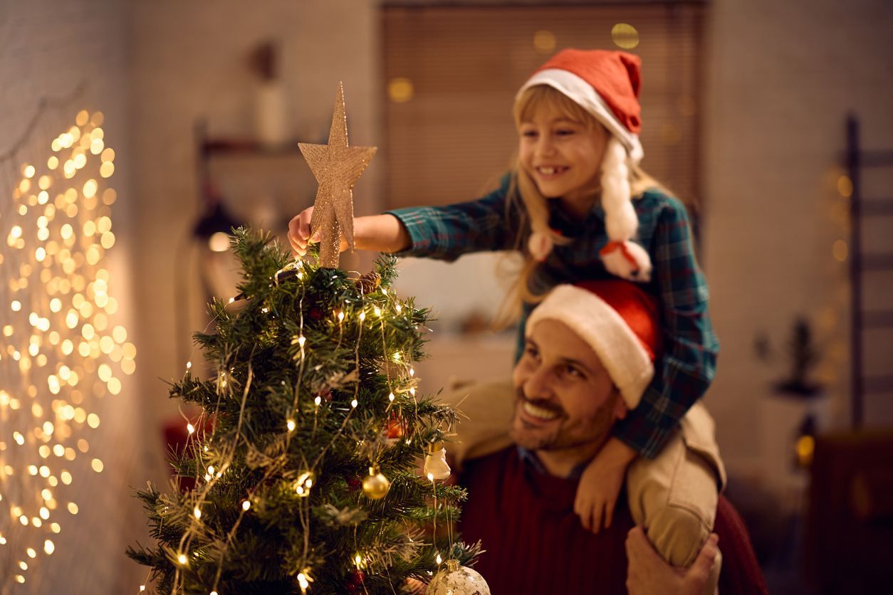 Girl in Santa hat places star on Christmas tree, perched on father's shoulders.