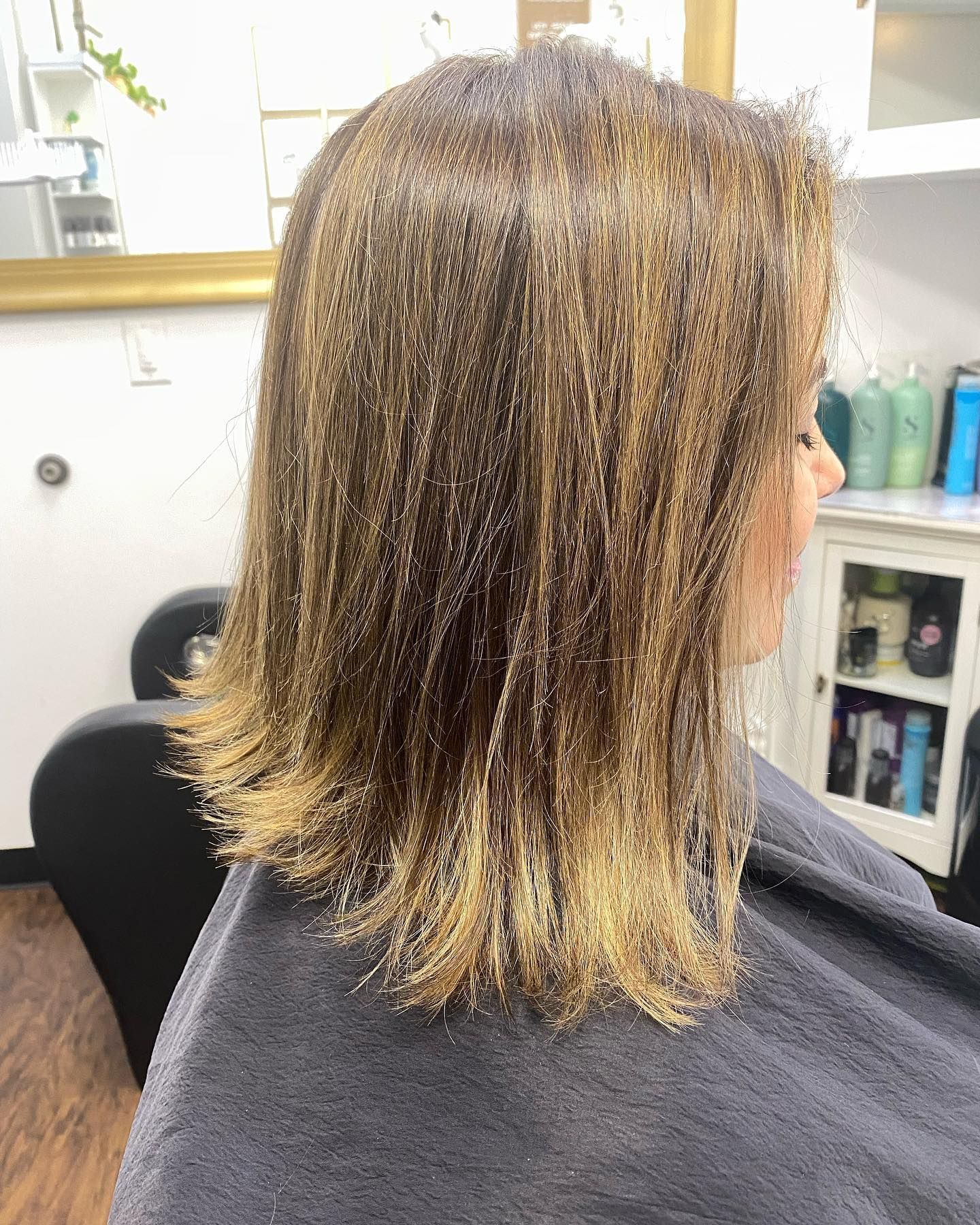 Woman with shoulder-length hair; light brown highlights, salon setting, seated in chair, wearing cape.