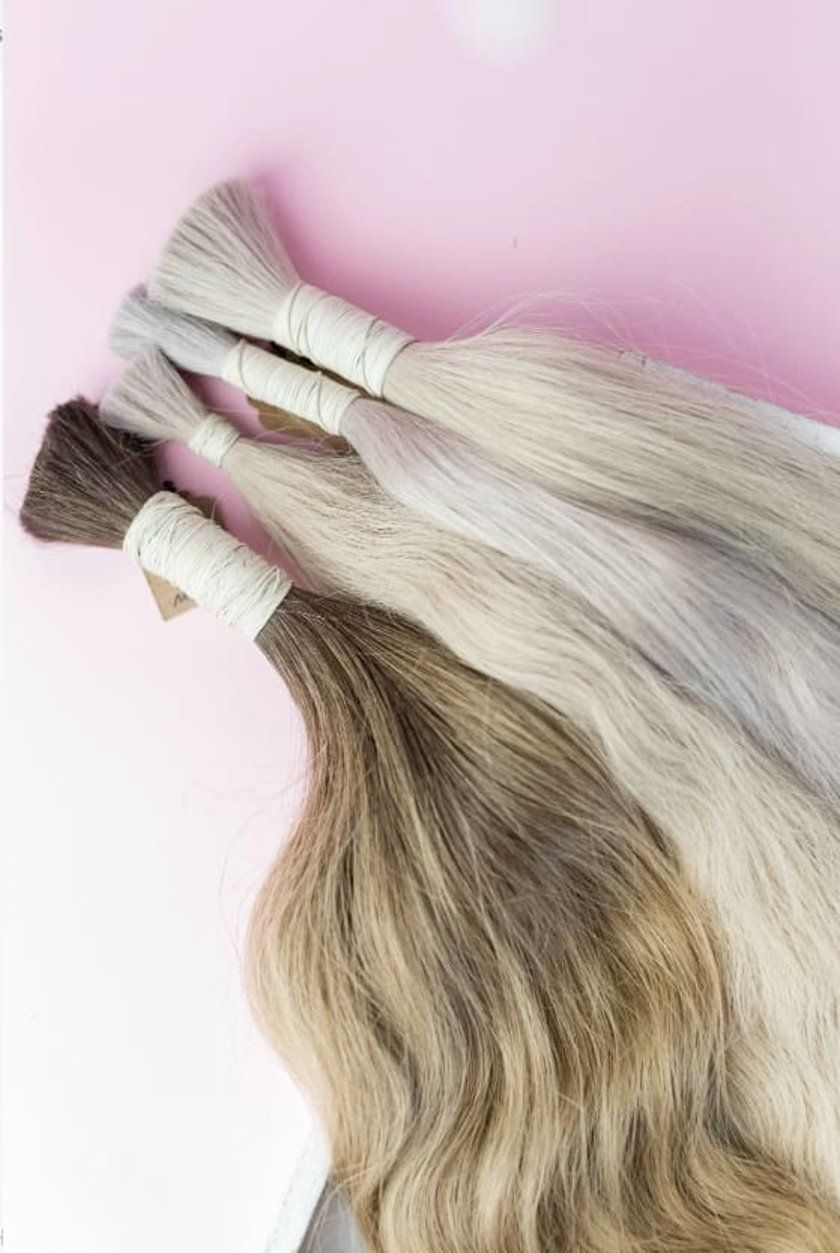 Bundles of long hair extensions in shades of brown, blonde, and gray, tied with white bands, on pink background.