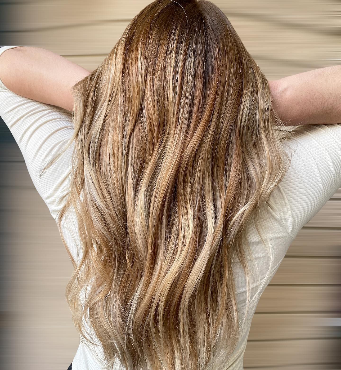Woman with long, wavy, highlighted brown hair. Arms behind head, wearing a white textured sweater.