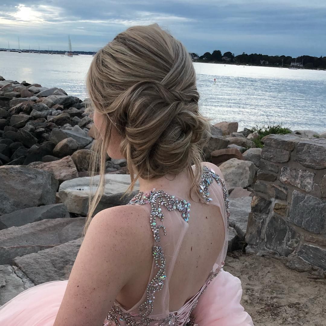 Woman in a pink dress with a decorated bodice, updo hairstyle, standing near rocks by the water.