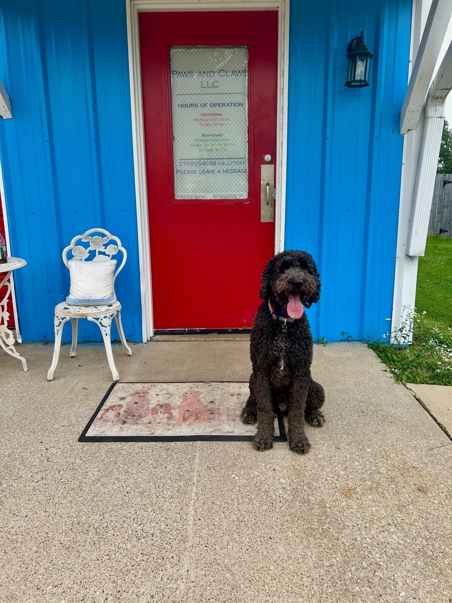 Black dog sits on a doormat in front of a red door and blue building.