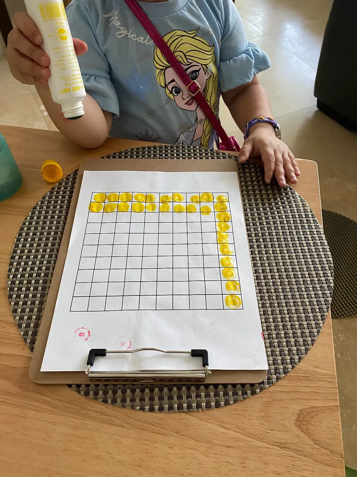 A little girl is sitting at a table with a clipboard and a bottle of glue.