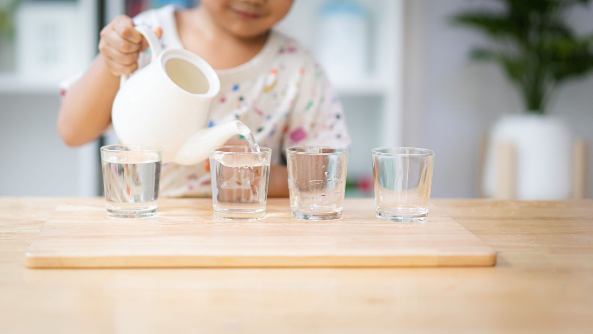 A little girl is pouring water into four glasses on a wooden table.
