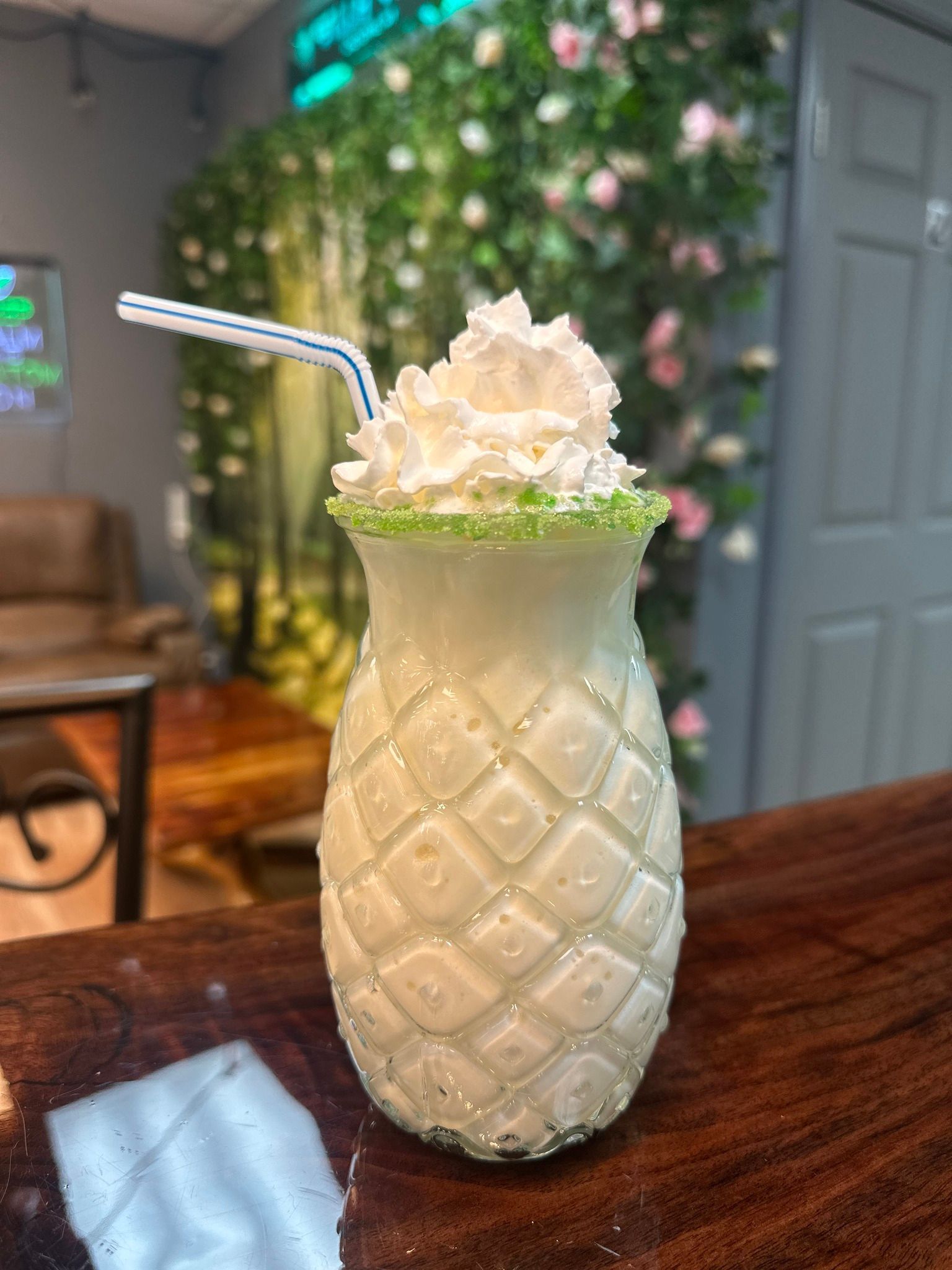 Pineapple-shaped glass filled with a light green drink, topped with whipped cream and a blue-striped straw, on a wooden counter.