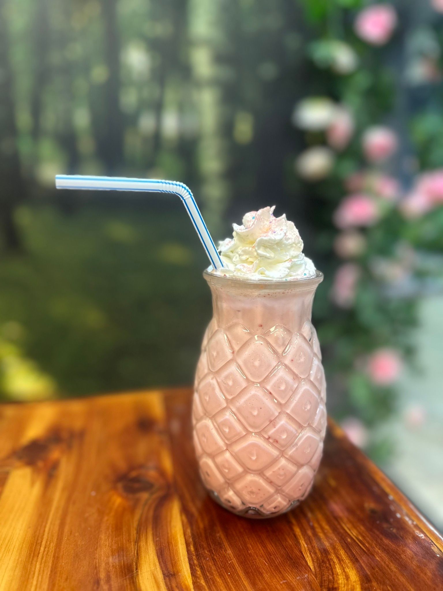 Pink pineapple-shaped milkshake with whipped cream and a straw on a wooden table, set against a blurry outdoor backdrop.