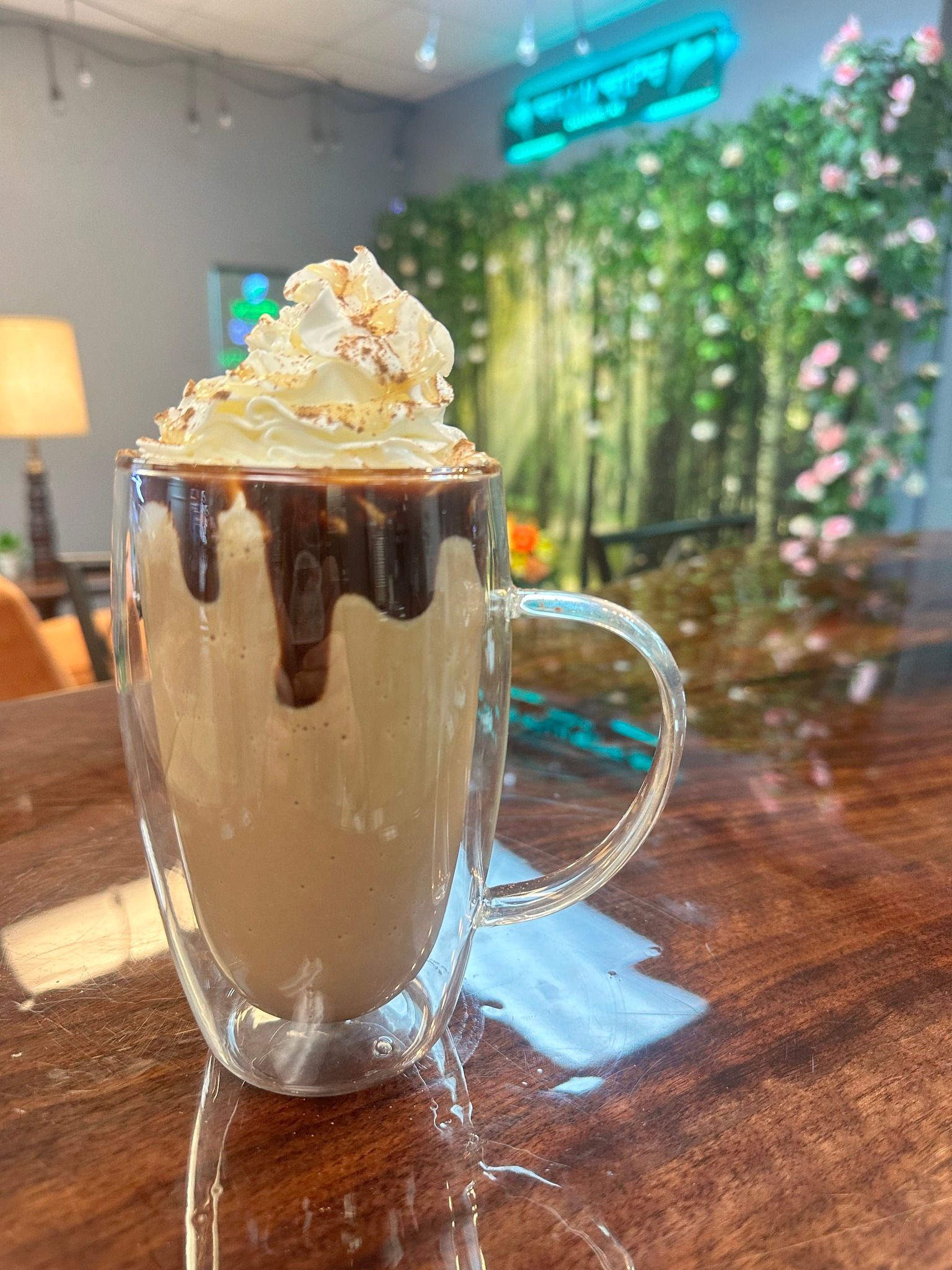 Hot drink in a clear glass mug, topped with whipped cream and chocolate drizzle, on a wooden table.