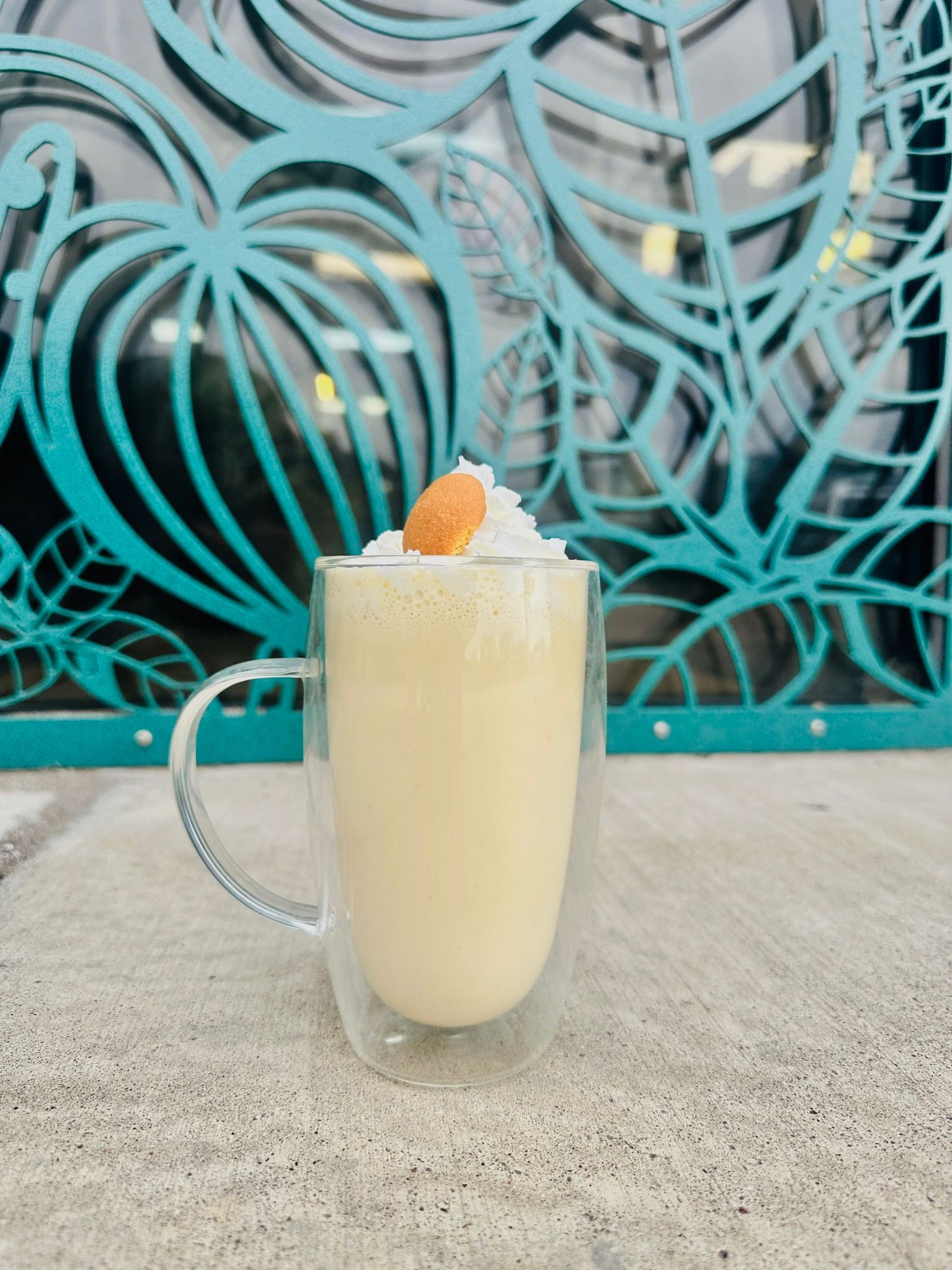 Banana pudding smoothie in clear mug, topped with whipped cream and wafer, against teal ornate background.