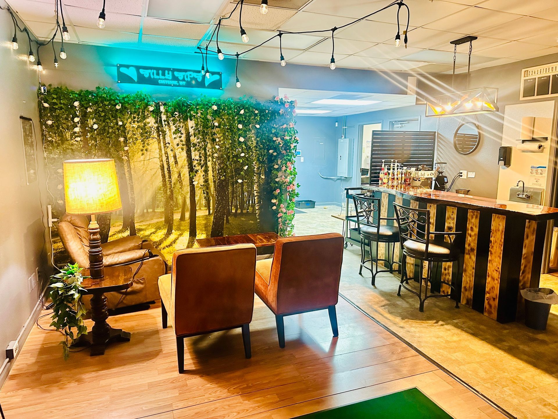 Cozy lounge with rustic bar, forest backdrop, and seating. String lights hang overhead.