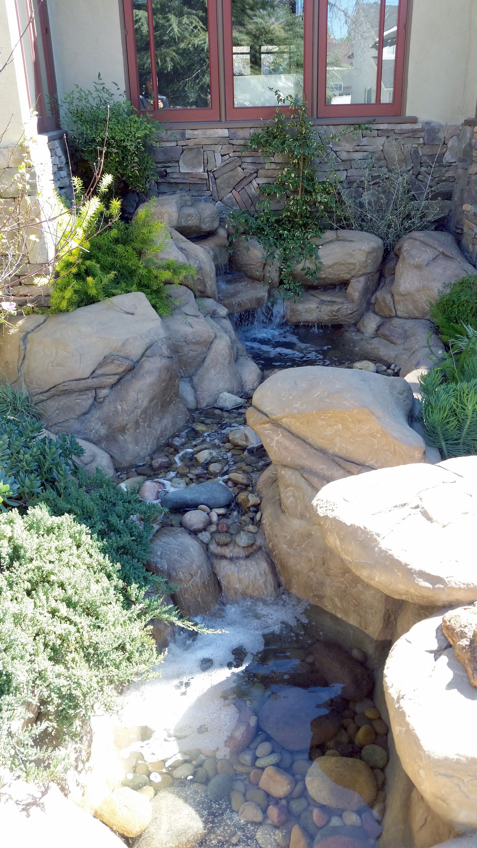 A small waterfall is surrounded by rocks and plants in front of a house.