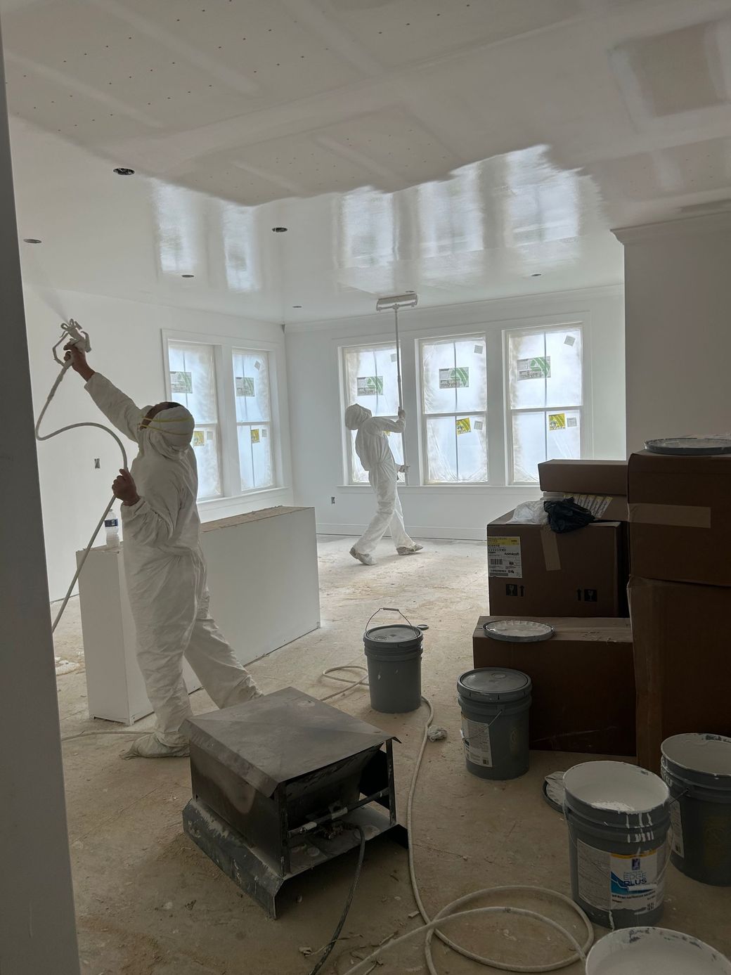 A man is spraying paint on the ceiling wearing a suit, and another man in a white suit is painting the ceiling with a roller.