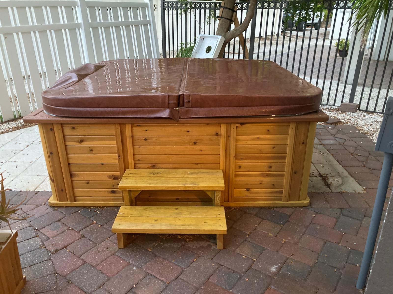 Hot tub with a brown cover, wooden exterior, and steps on a brick patio.