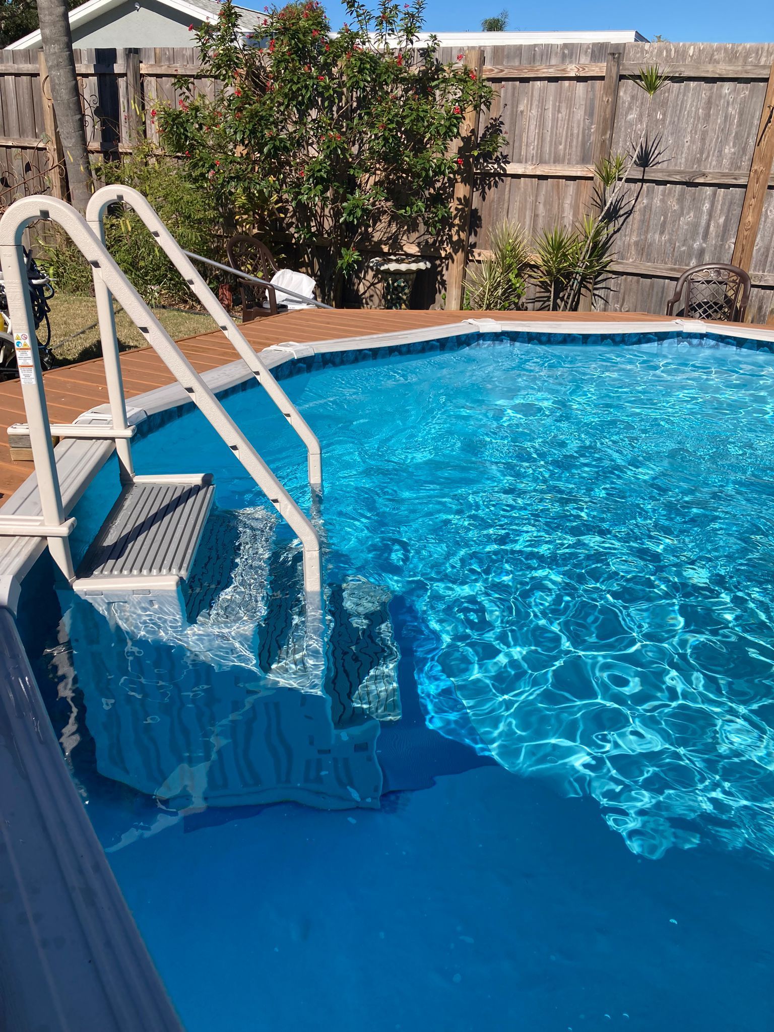 Above-ground pool with built-in stairs; blue water, white ladder, brown deck, wooden fence.
