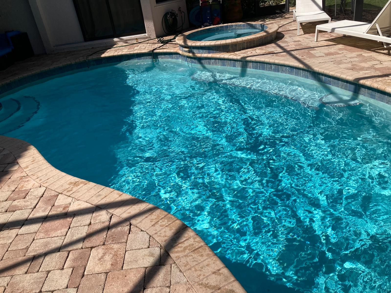 Outdoor swimming pool with clear blue water and brick patio. Hot tub in the background.