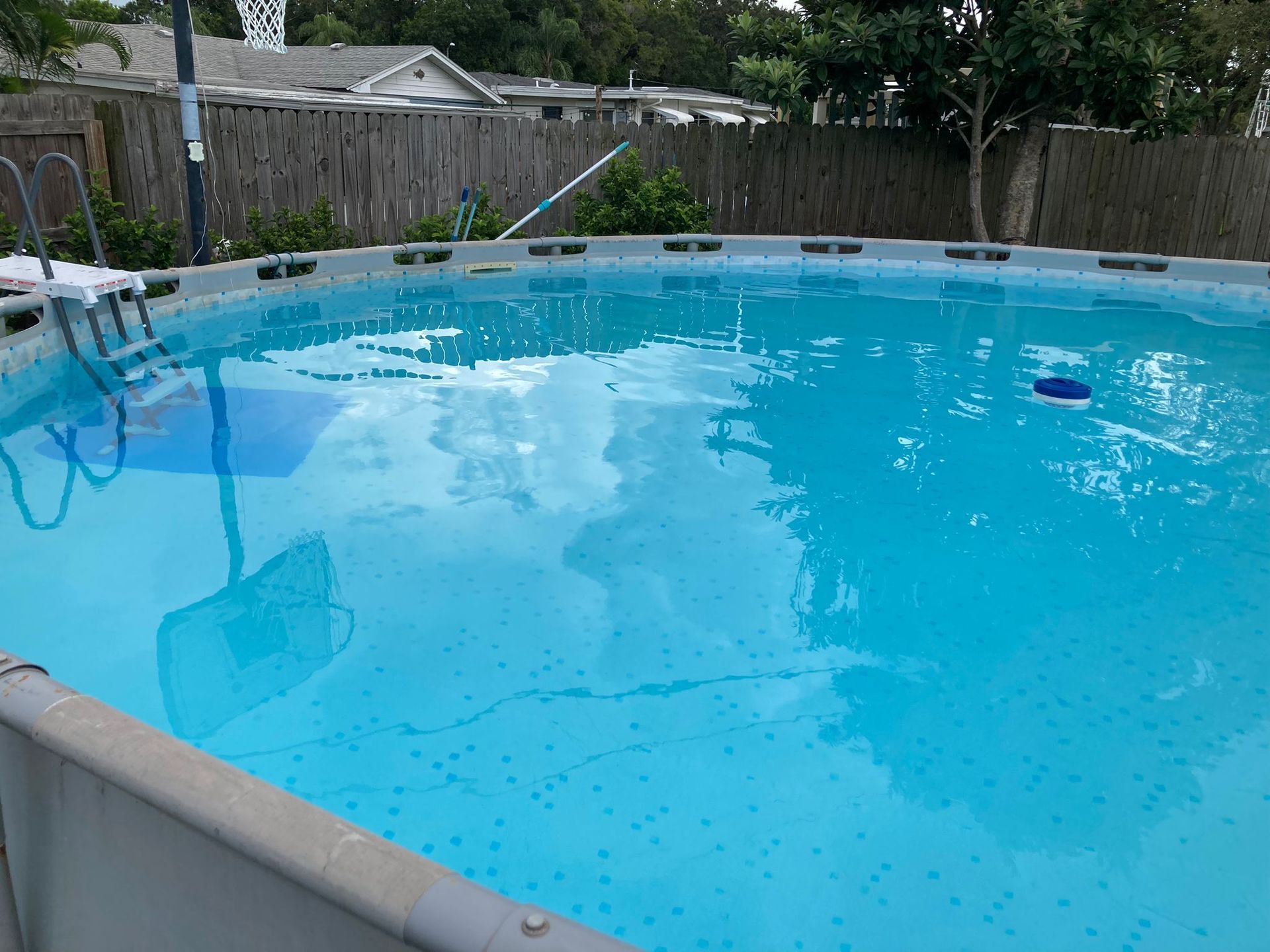 Above-ground pool with clear blue water, a ladder, and basketball hoop in a backyard setting.