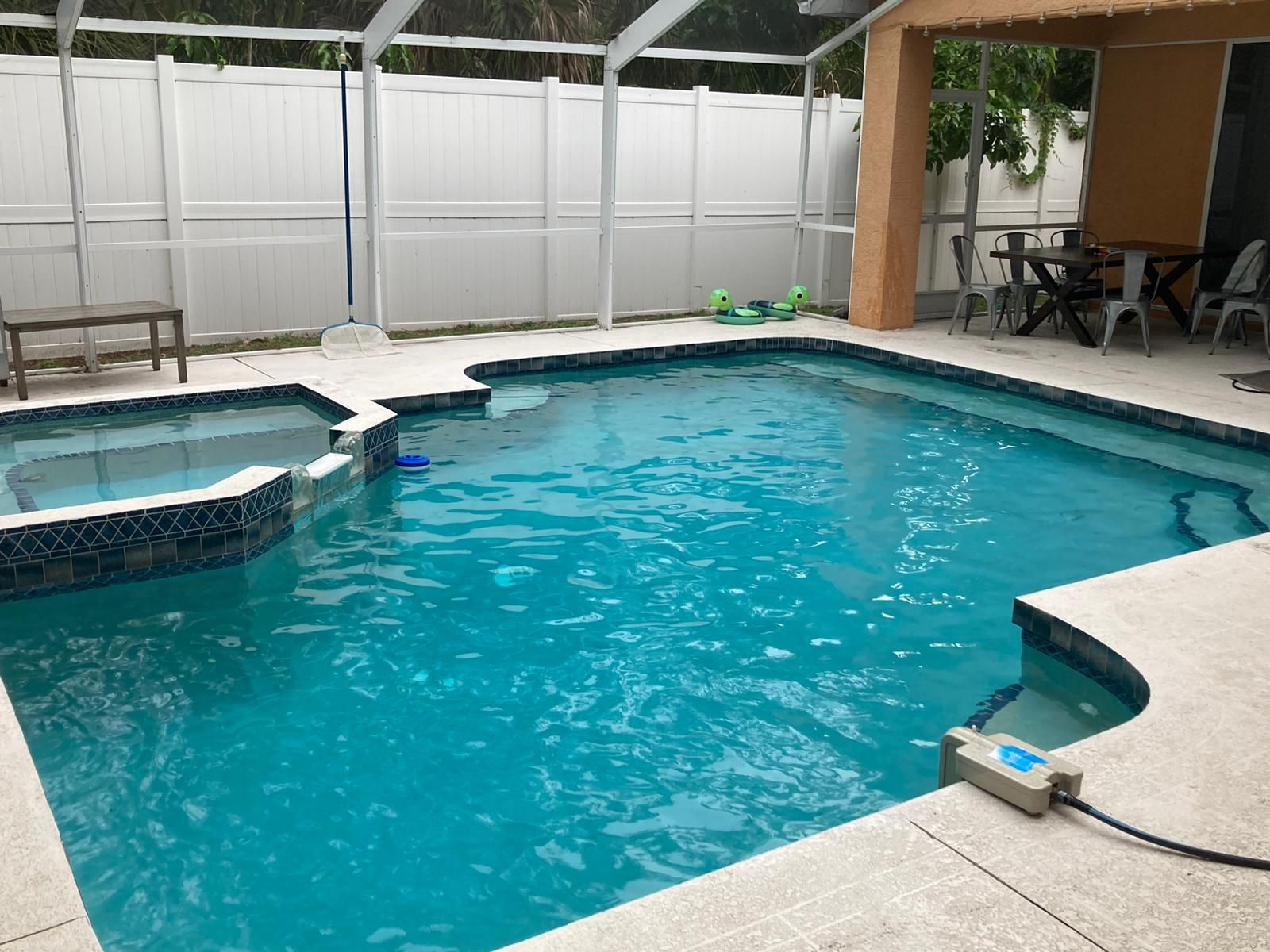 Pool with turquoise water, jacuzzi, patio furniture, and white fence in a screened outdoor area.
