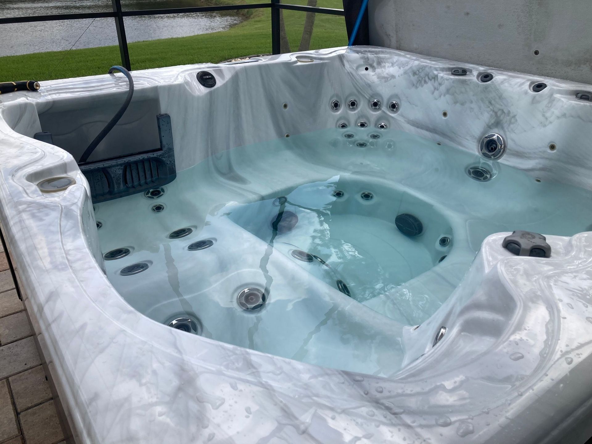 Hot tub with clear water, surrounded by grey and white marbled surface, outdoors with a pond in the background.