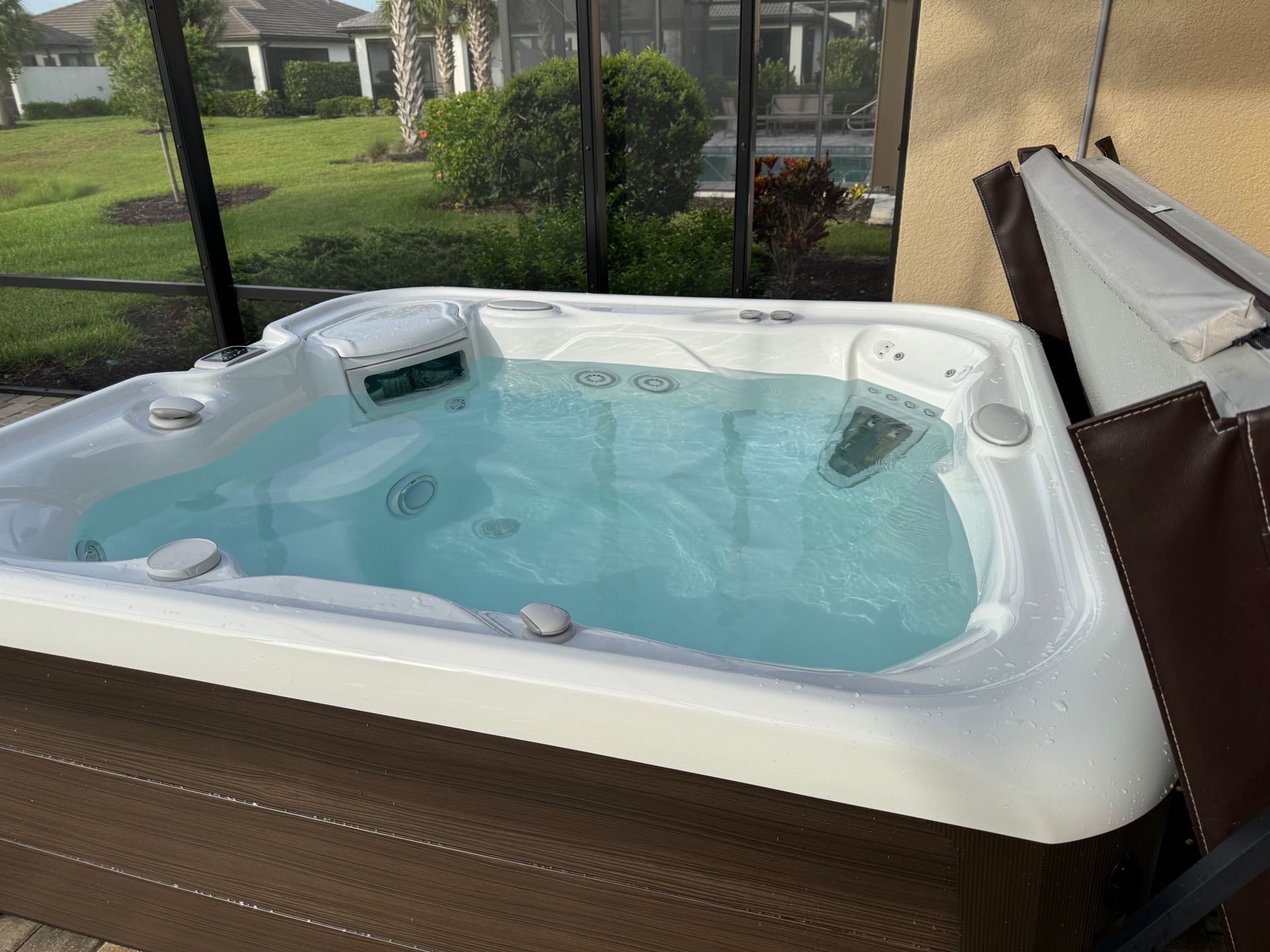 A white hot tub with clear water sits outdoor. The partially opened brown cover is to the right.