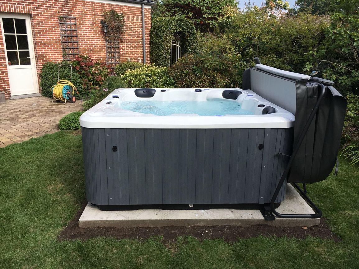 Hot tub in a backyard setting, surrounded by grass and a brick building. Cover is open.