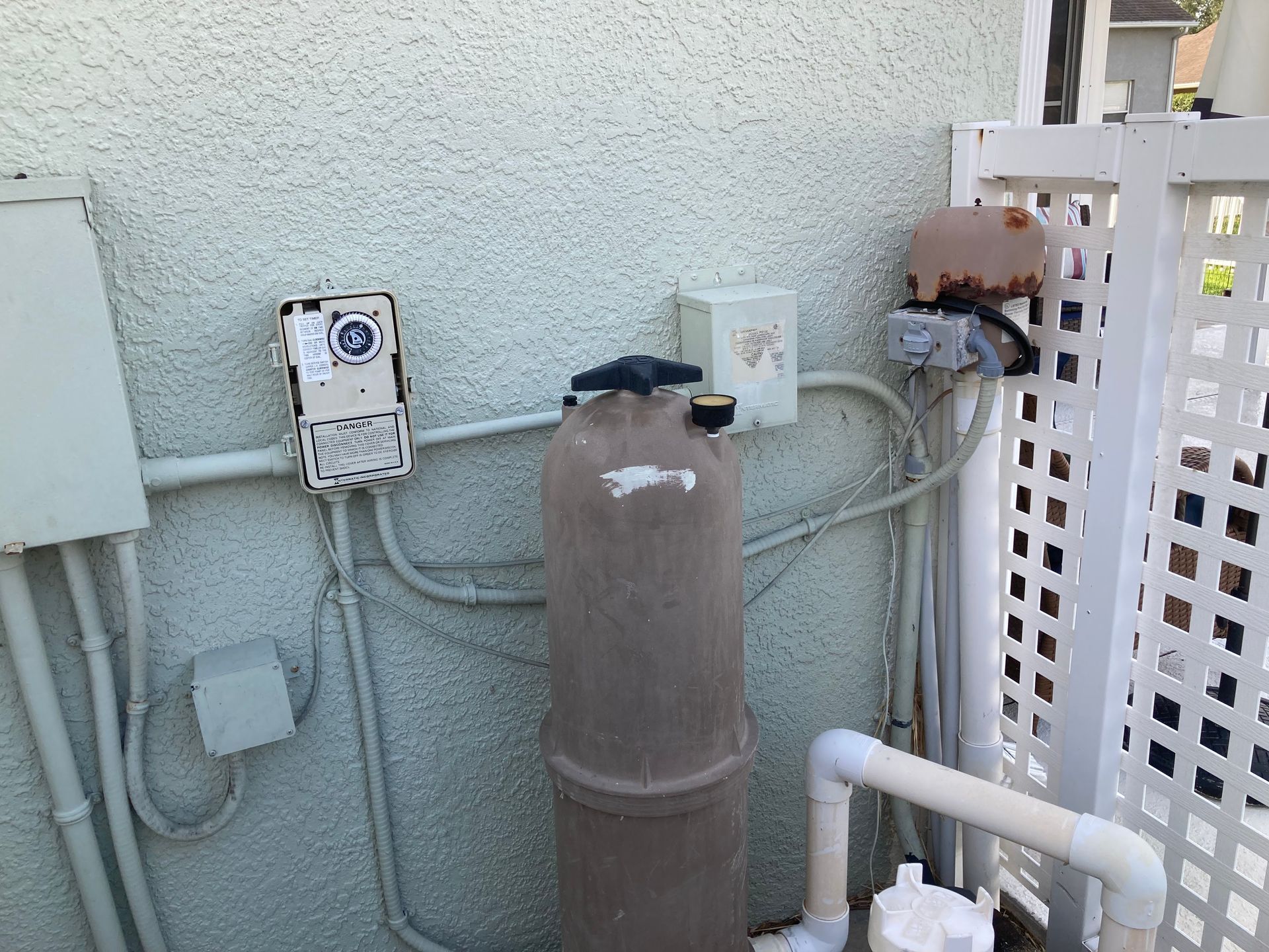 Pool equipment against a textured wall and white fence. Includes a filter, electrical boxes, and plumbing.