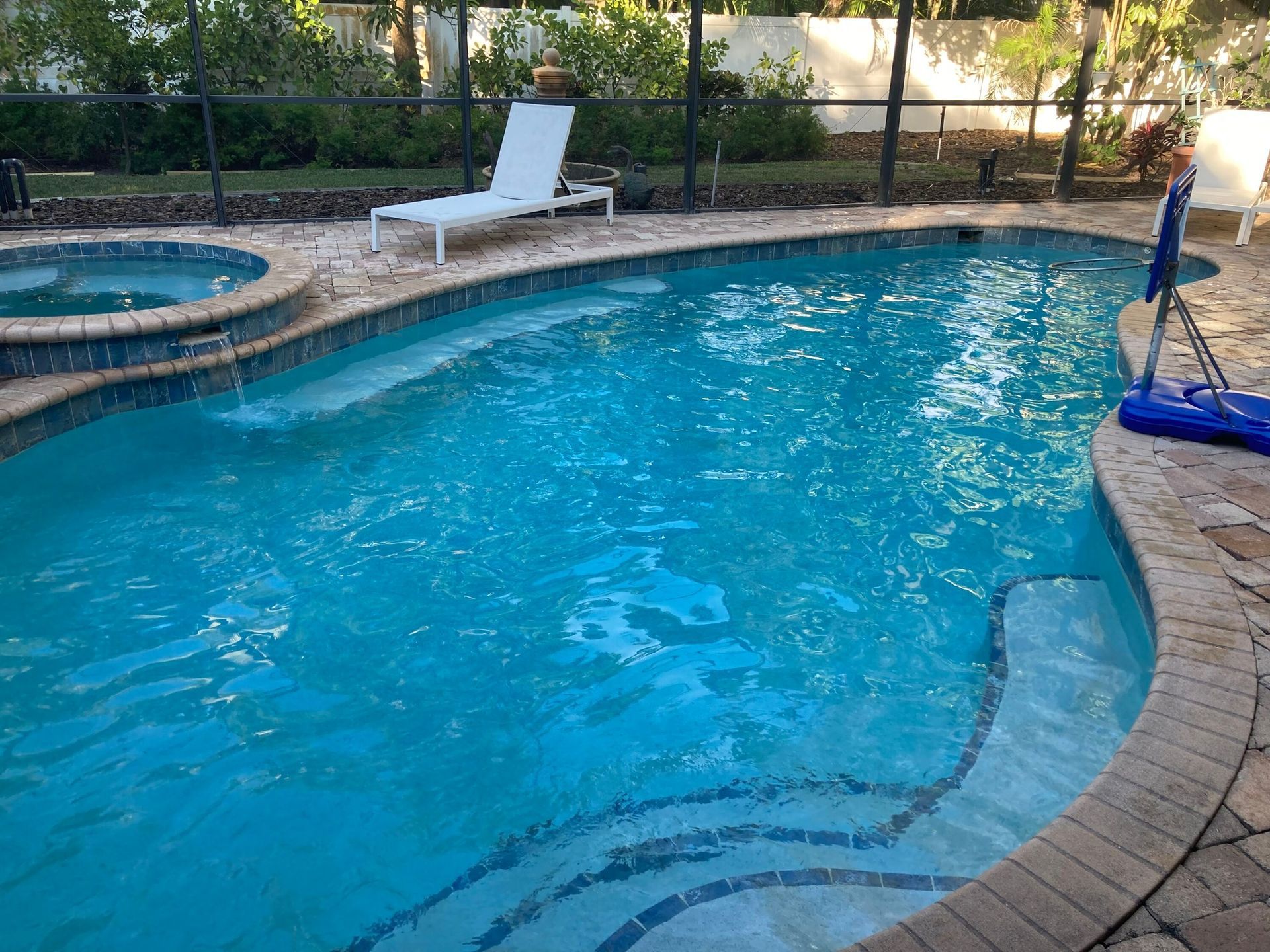 Swimming pool with attached jacuzzi.  Blue water, stone deck, white lounge chair, basketball hoop, and black fence.