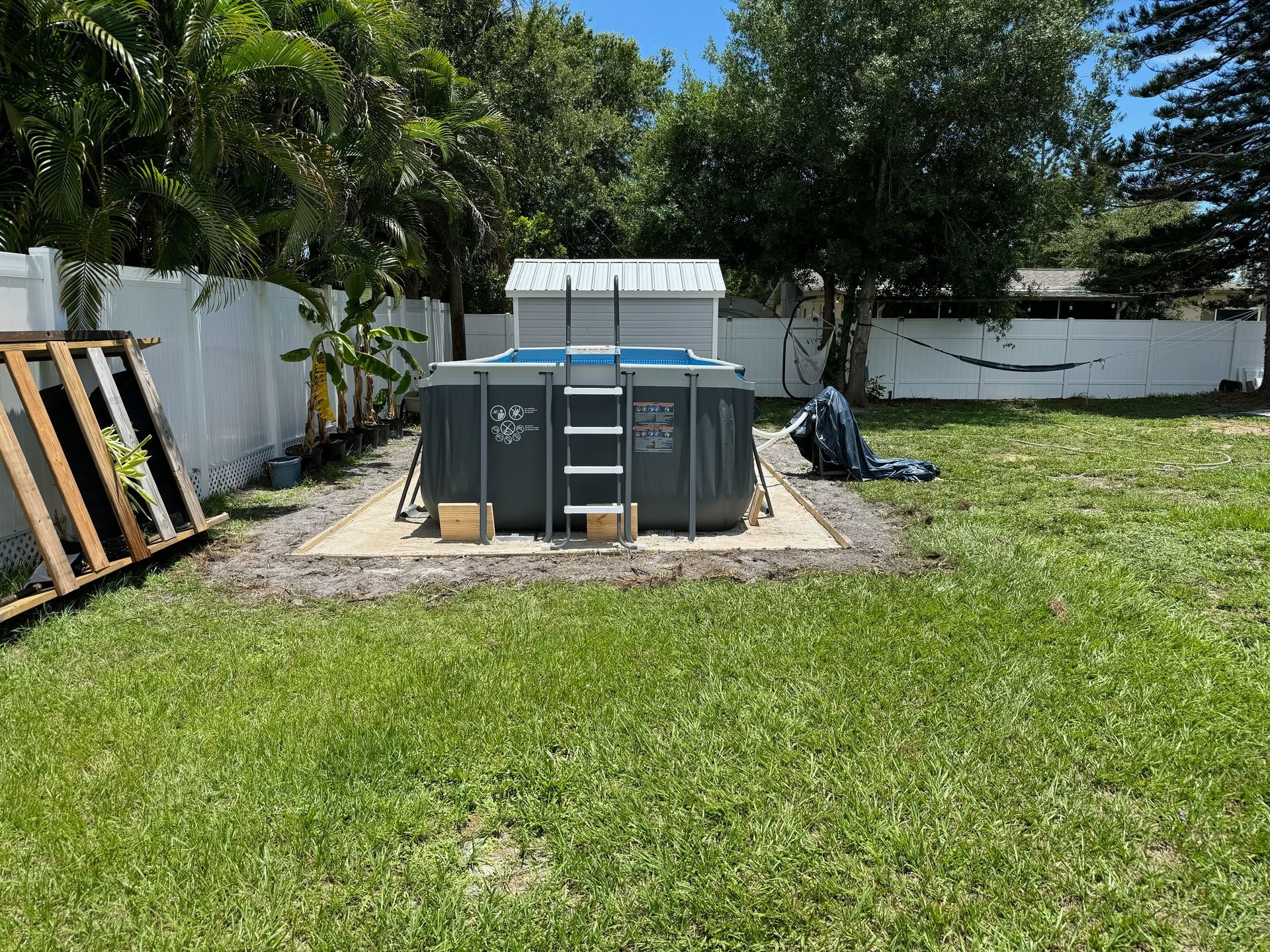 Backyard with an above-ground pool on a concrete pad, surrounded by a white fence and greenery.