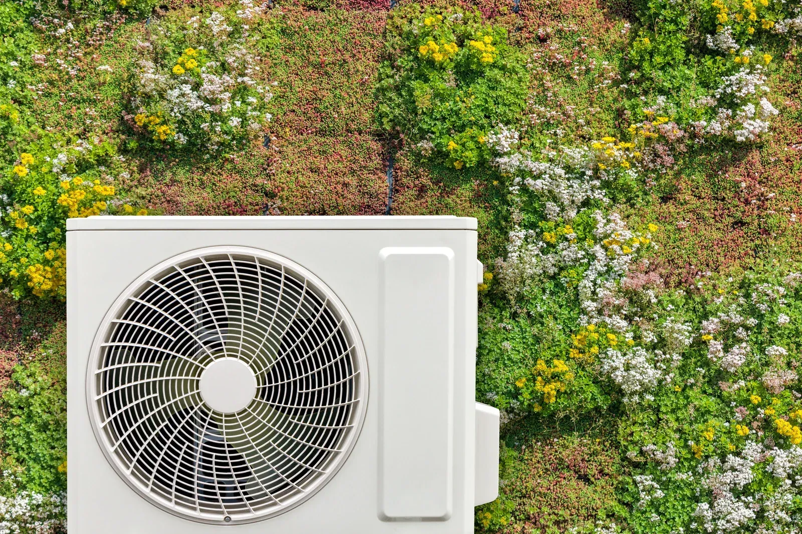 White air conditioner unit against a green wall with various plants and flowers.