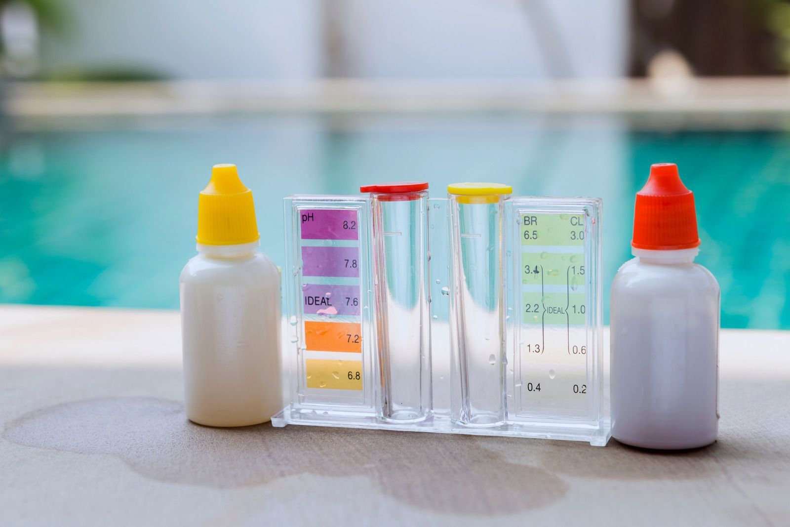 Pool water test kit with vials and color chart next to white bottles, pool in background.