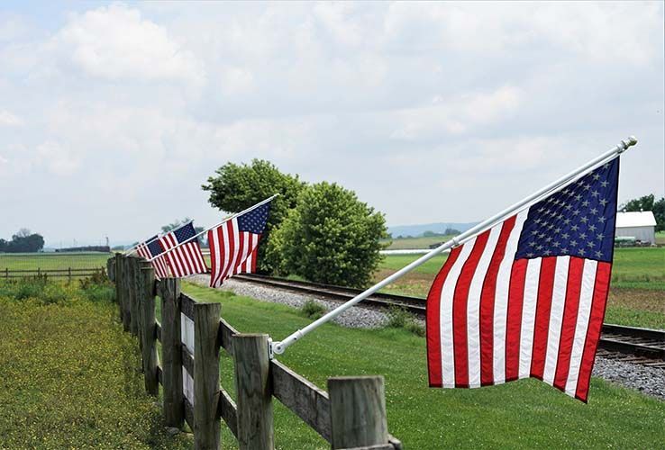 American flags on a wooden fence, railway track, and farmland under a cloudy sky.