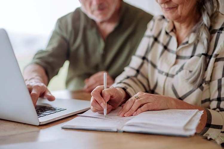 Man and woman working on a laptop, woman writing in a notebook.