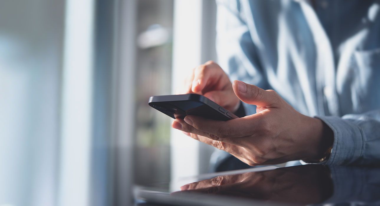 Person's hands holding a smartphone, tapping the screen, near a window, natural light.