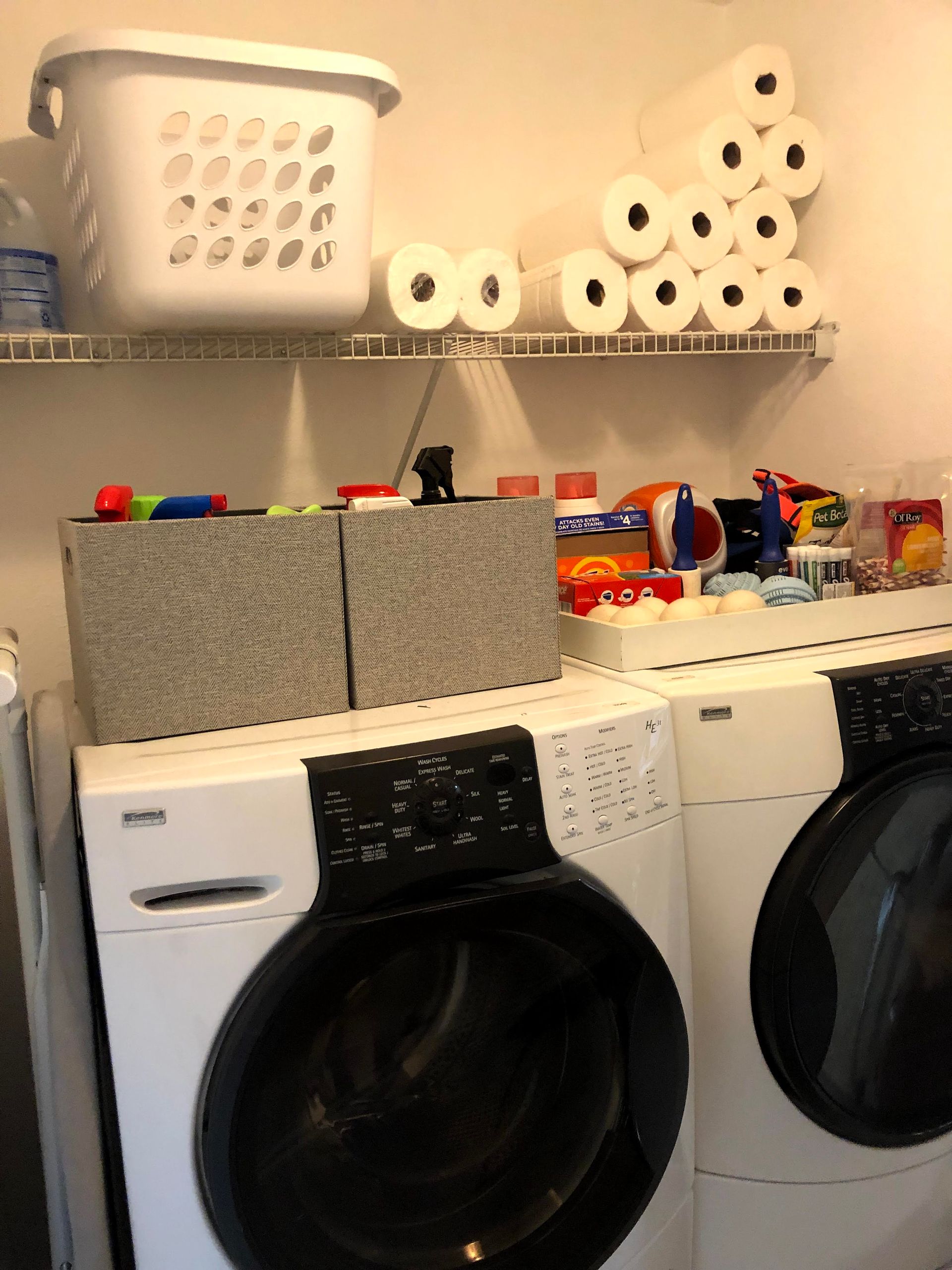 Laundry room with white washer and dryer, laundry basket, and shelves with paper towels and supplies.