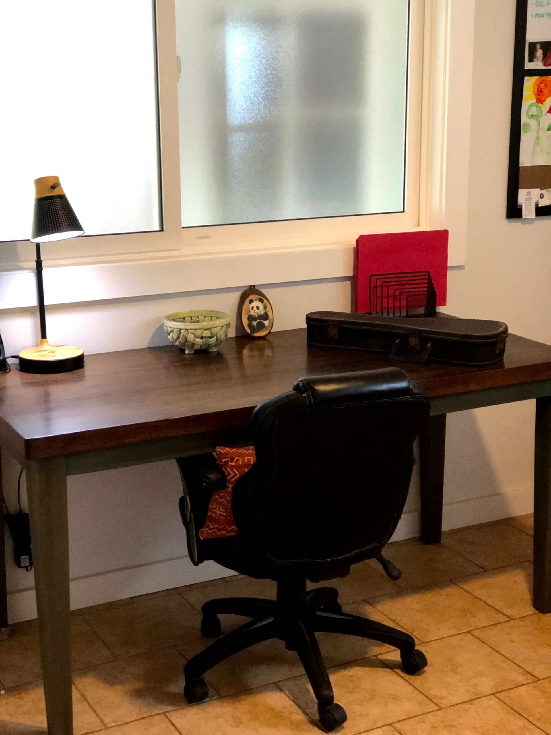Desk with black chair, lamp, and items on top, in front of a window.